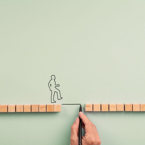 Education, mentorship and support conceptual image Male hand drawing a bridge between two rows of wooden blocks for a silhouetted man to walk across. Over blue background with plenty of copy space.